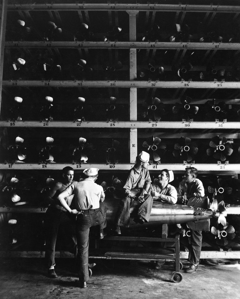 Group of torpedomen relaxing beneath rows of deadly torpedoes in torpedo shop, one man actually sitting on torpedo resting on gurney, during WWII. (Photo by Horace Bristol/The LIFE Picture Collection © Meredith Corporation)