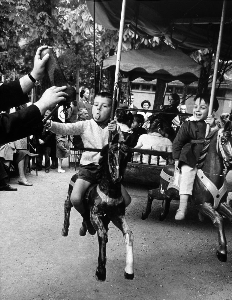 Little boy on merry-go-round at the Tuileries Gardens, sticking out his tongue, 1963.